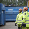 Two recycling experts dressed in hardhats and reflective Stena Recycling work clothes standing beside a couple of large blue recycling containers in a recycling yard.